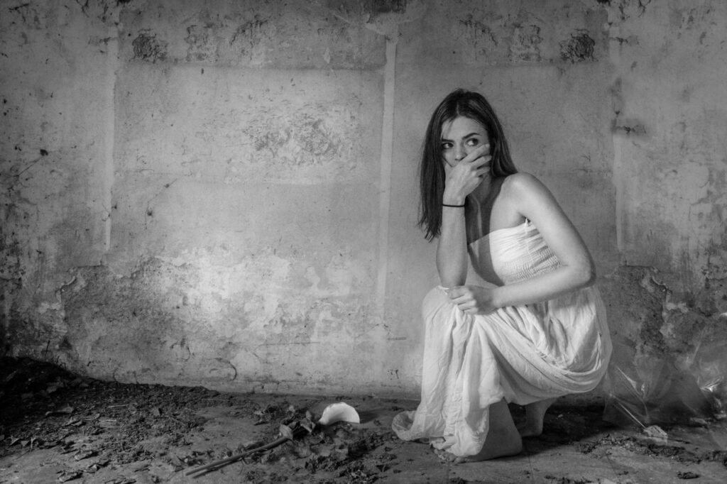 A woman in a white dress sits thoughtfully in an abandoned room, highlighting decay.